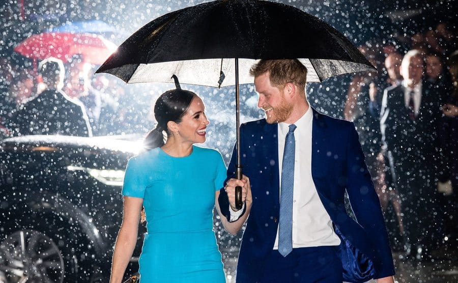 Meghan Markle and Prince Harry smiling at each other under an umbrella in the rain with a light behind them making the rain light up around them 