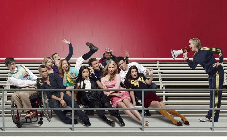 The cast of Glee sitting on bleachers being blown to the side by Jane Lynch yelling into a microphone 