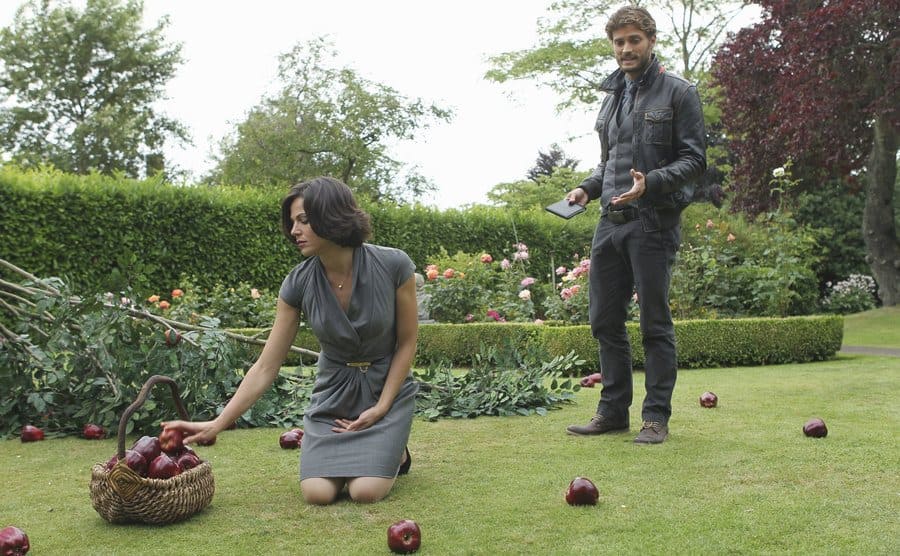 Lana Parrilla picking up apples and placing them in a wicker basket while Jamie Dornan stand behind her explaining something in a garden 