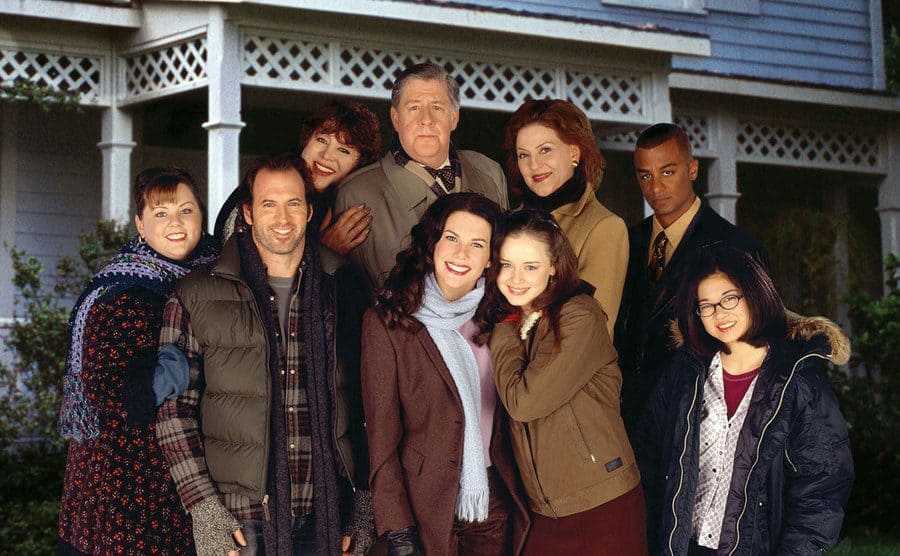 The cast of Gilmore Girls posing outside of Lorelai and Rory’s old white house 