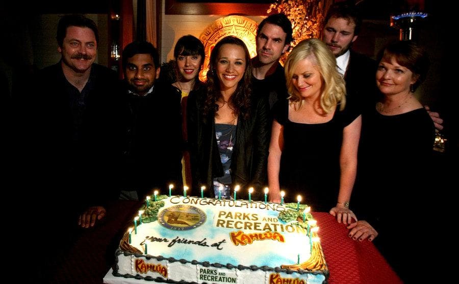 The cast of Parks and Recreation posing around a cake celebrating the show’s premiere 
