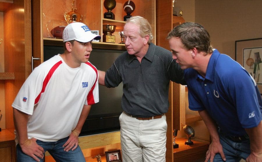 Eli, Archive, and Peyton Manning huddling in front of a plasma tv surrounded by family photographs and trophies