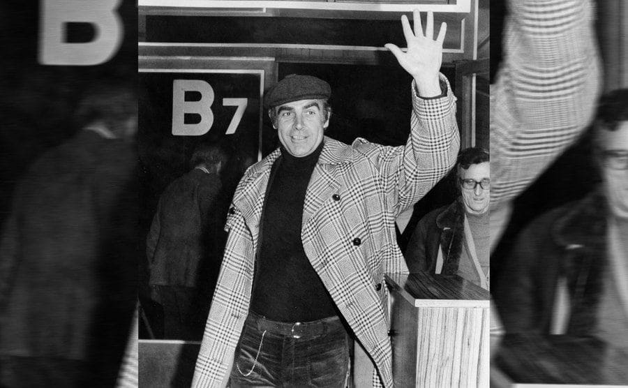 Sean Connery waving while walking with his rolling suitcase at the airport 