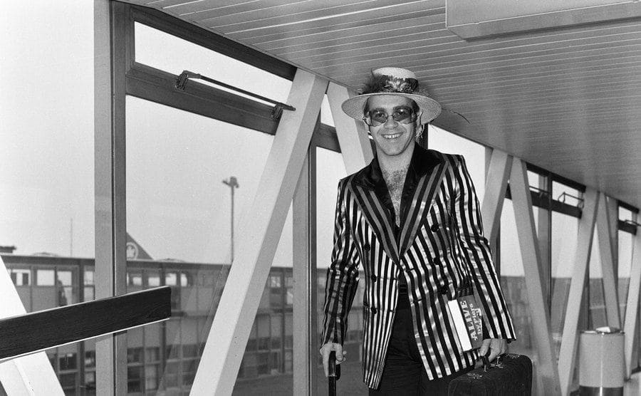 Elton John walking down a hallway in the airport with a striped suit on 
