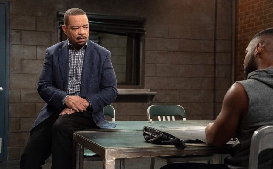 Ice T as Fin Tutuola sitting on the edge of a desk in the interrogation room with a suspect sitting across from him 