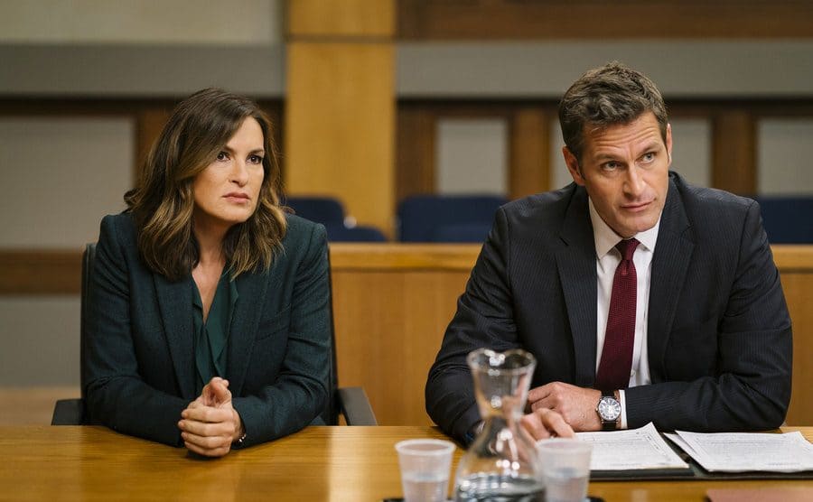 Mariska Hargitay and Peter Hermann sitting behind the defenses desk in a courtroom in a scene from Law and Order