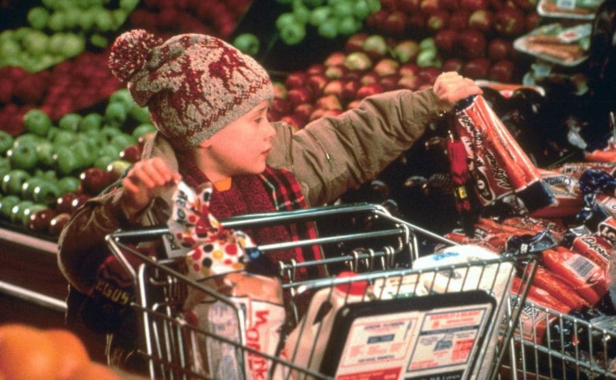 Macaulay Culkin pushing a shopping cart with wonder bread and milk inside while picking up a bag of carrots in the refrigerated vegetable section 