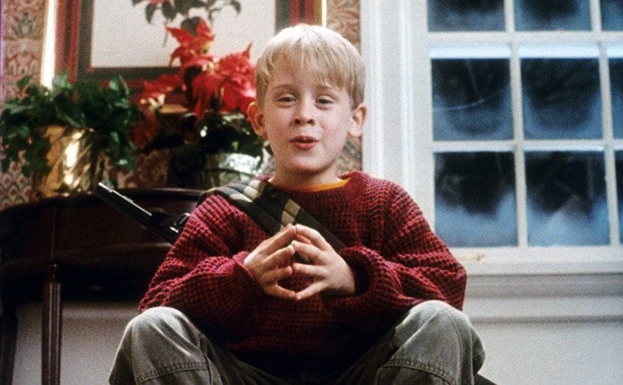 Macaulay Culkin sitting with his fingers together in the front entrance of the house in Home Alone 