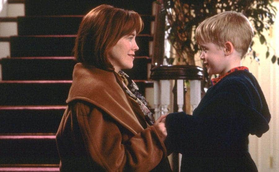 Catherine O’Hara holding Macaulay Culkin’s hands kneeling next to him in front of a large staircase 