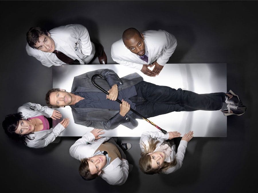 The cast of House M.D. is standing around a hospital bed while Greg House is on the bed, promotional photo. 