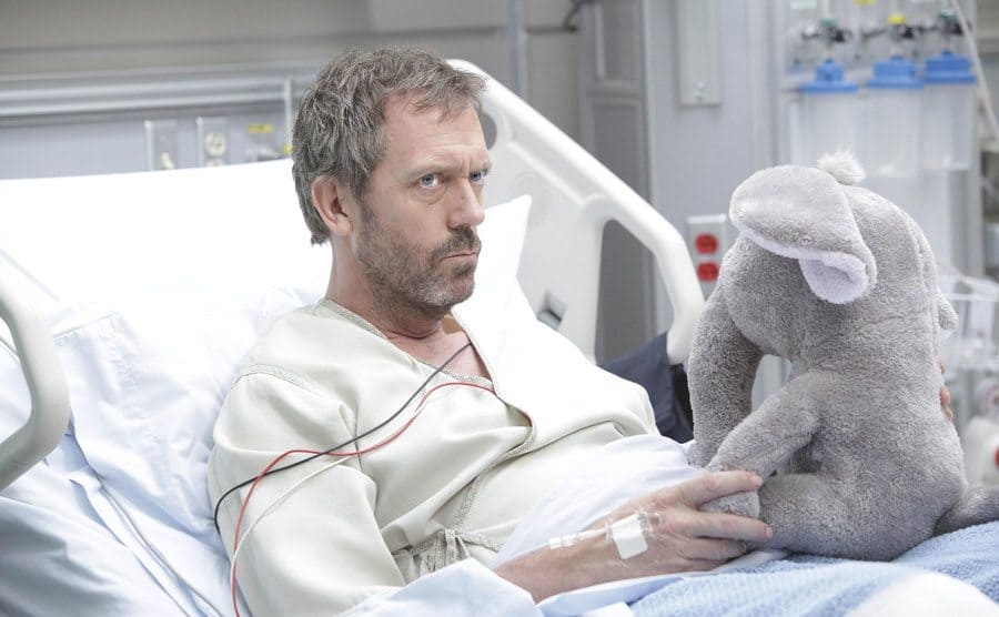 Greg House is lying in a hospital bed with a stuffed elephant doll in his hands. 