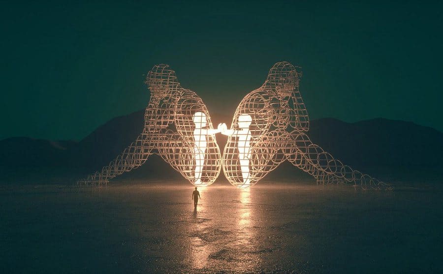 Two metal figures holding hands lit up inside of two metal outlines of adults sitting with their backs towards each other with a man walking towards it 