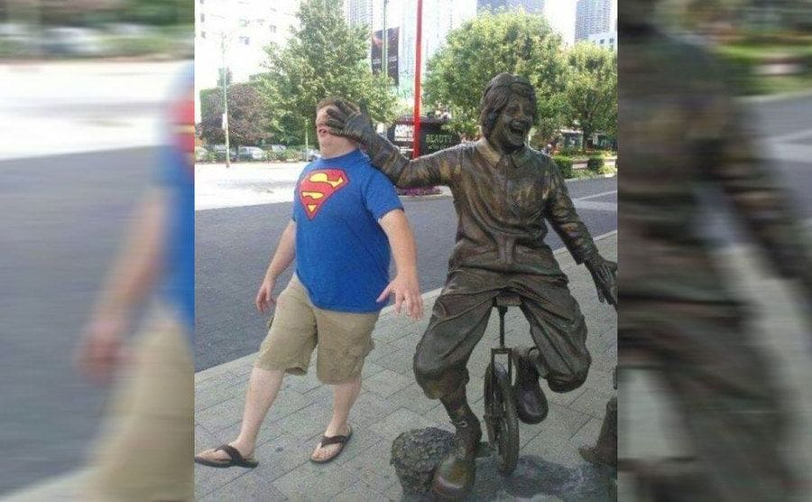 A guy wearing a superman shirt walking into the hand of a statue of a man riding a unicycle 