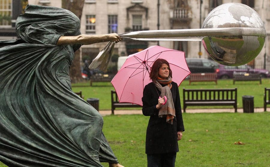 A woman with a pink umbrella standing next to the statue of a woman seeming to pull the earth towards her 