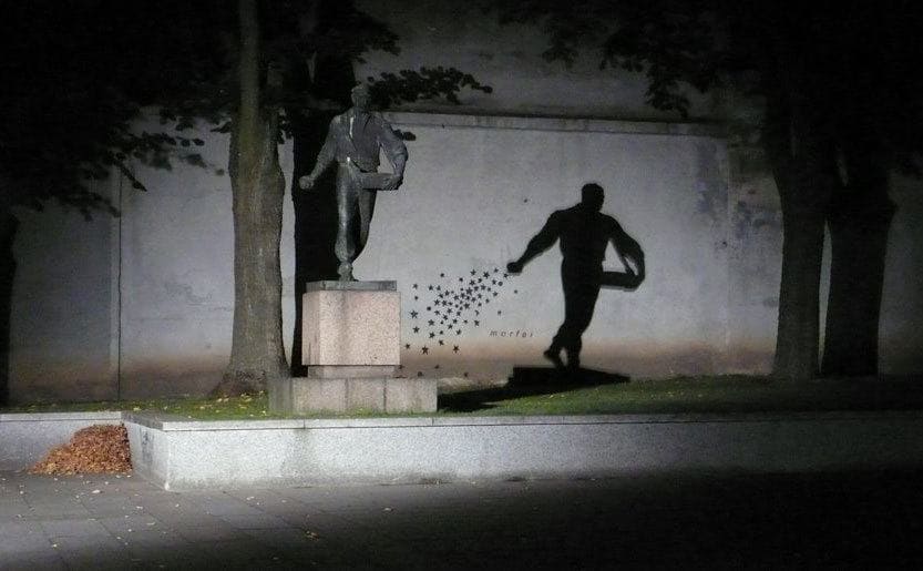 A statue of a man with his hand in a fist holding a box with his other hand, and stars painted on the wall behind him giving the illusion of him sprinkling the stars when the light hits it right 