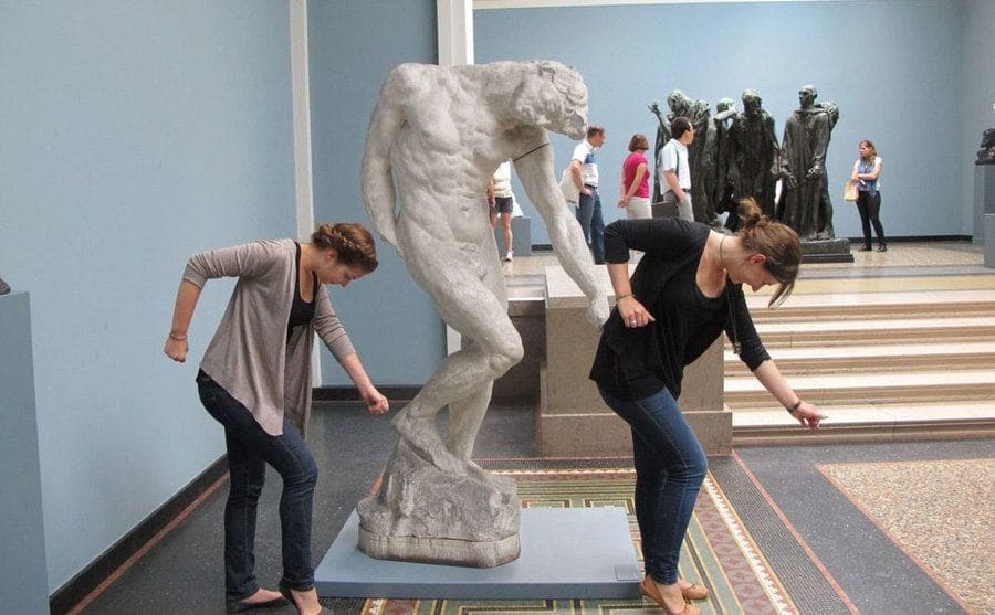 Two women next to a statue posing like it in a museum 