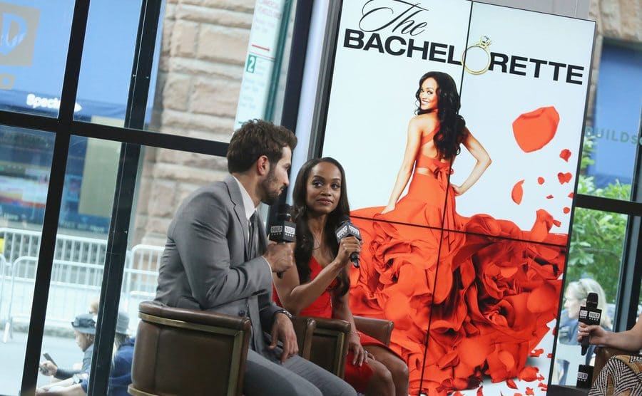 Bryan Abasolo and Rachel Lindsay on a talk show with an ad for The Bachelorette behind them
