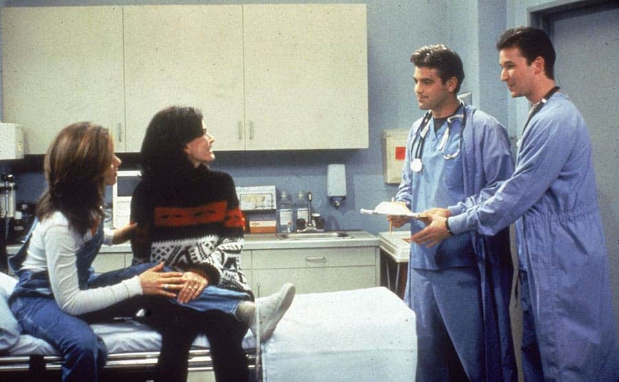 Jennifer Aniston and Courteney Cox sitting in a hospital bed with George Clooney and Noah Wyle as doctors 