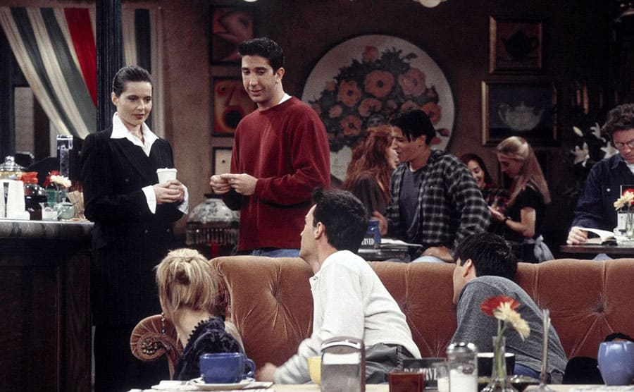 Isabella Rossellini standing with David Schwimmer at the bar getting coffee while Lisa Kudrow, Matthew Perry, and Matt LeBlanc hide behind the couch watching them 