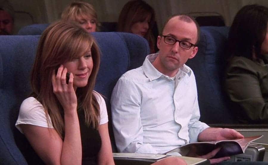 Jim Rash looking at Rachel on the phone while sitting on an airplane 