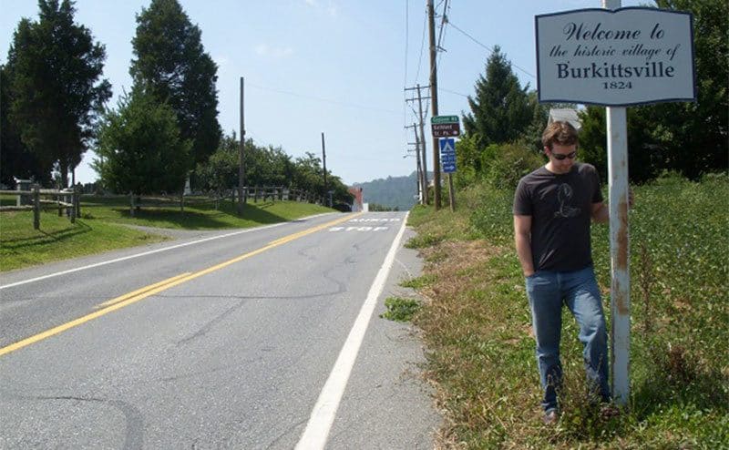 A man is standing next to the Burkittsville sign posing for a photo.