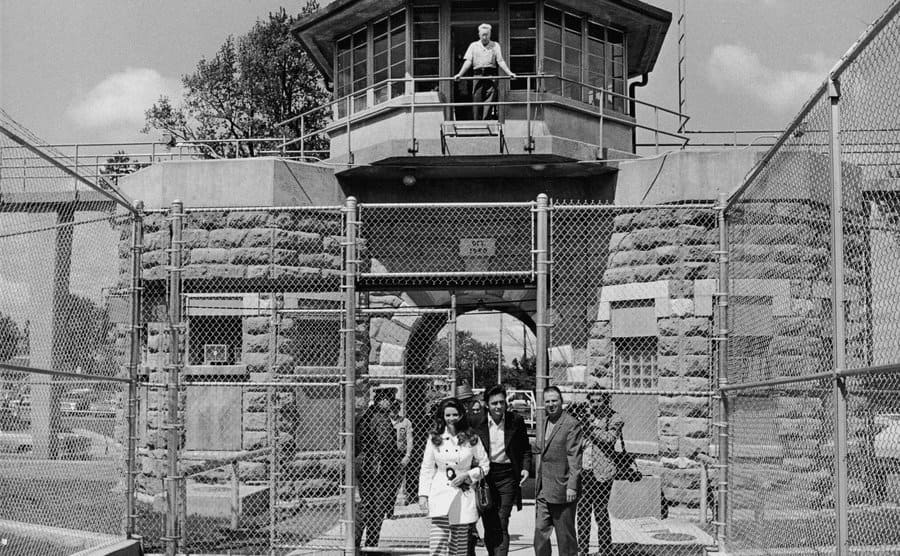 June Carter and Johnny Cash walking out of a prison entrance with guards around them and another guard standing in the guard tower above them 