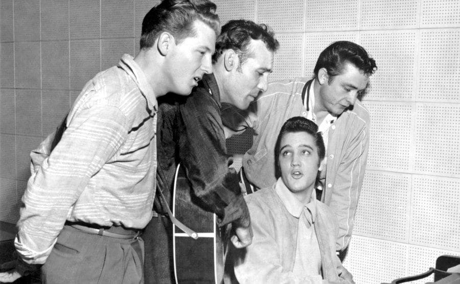 Johnny Cash standing with Jerry Lee Lewis, and Carl Perkins around Elvis Presley, who is sitting at a piano playing