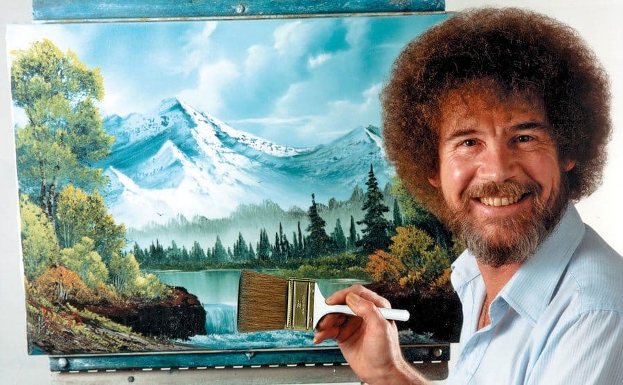 Bob Ross holding a large paintbrush in front of a mural 