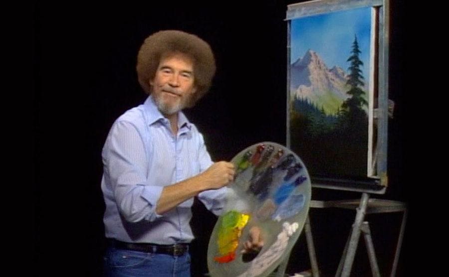 Bob Ross with a platter of paint in front of an easel 