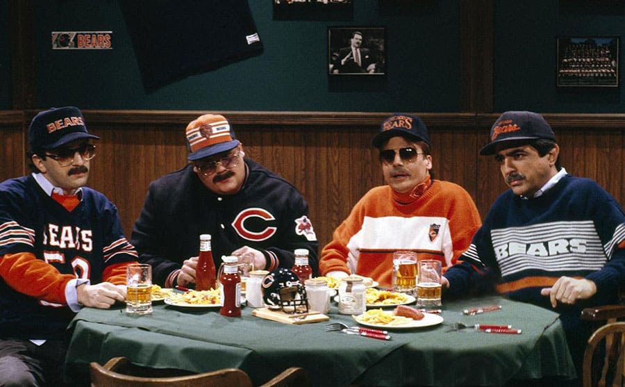 Robert Smigel, Chris Farley, Mike Myers, and Joe Mantegna sitting around a sports bar in Bears hats and uniforms in a scene from SNL