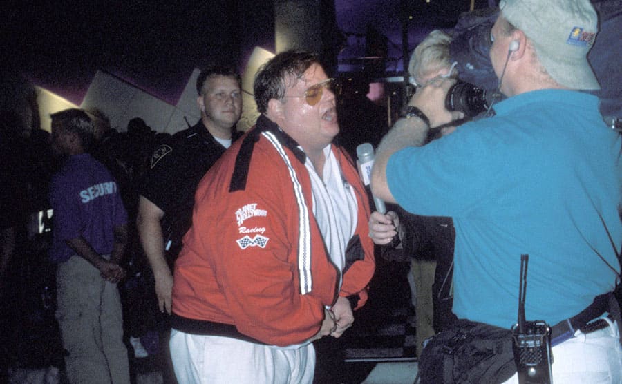 Chris Farley acting intoxicated in front of the press while an officer stands by 
