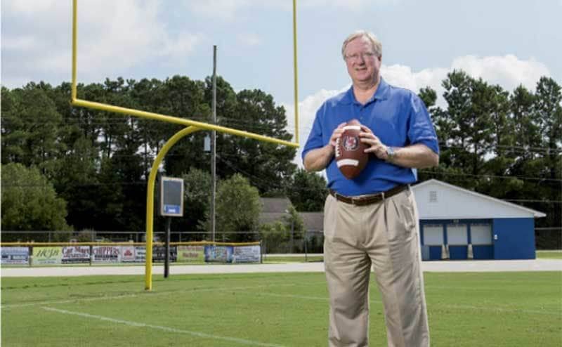 an older Ronnie Bass stands on a football field holding a football in his hands. 