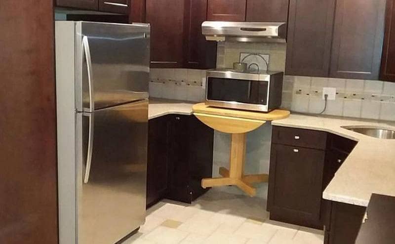 A microwave placed over a table in the empty space in the kitchen where typically an oven would be. 