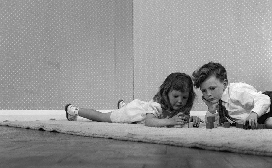 A girl and a boy playing with wooden building bricks on a rug, circa 1960.