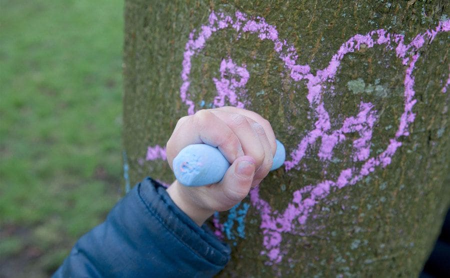 Child drawing a heart on a tree trunk with chalk.
