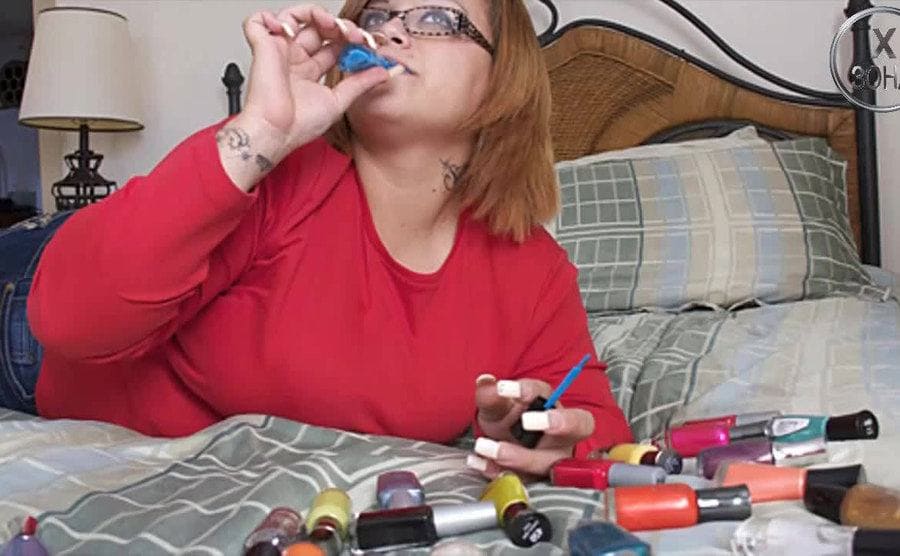 Bertha on her bed surrounded by nail polish bottles as she drinks from a blue color nail polish.