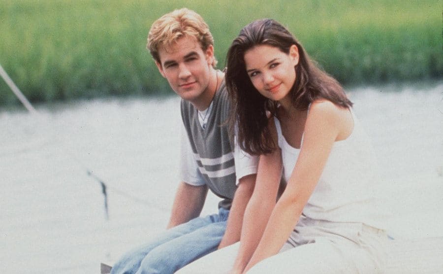 James Van Der Beek and Katie Holmes sitting on the edge of the patio which stretches out over the lake behind them 