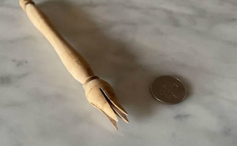 A wooden four-prong olive pick on a table next to a penny for scale 