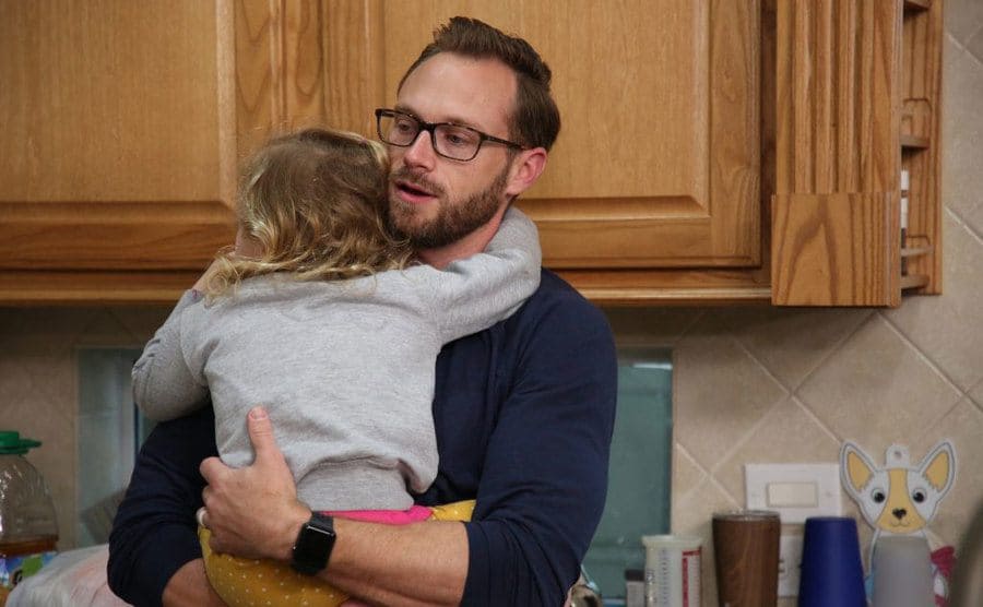 Adam is standing in the kitchen as he holds his daughter in his arms. 
