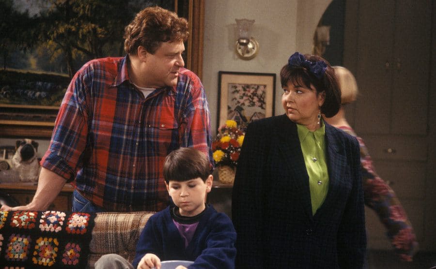 John Goodman and Roseanne standing around the couch 