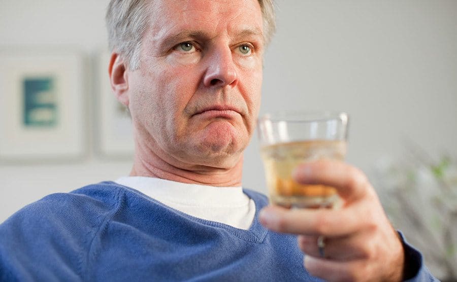A man is looking rather unhappy holding up a glass as he sits at home. 
