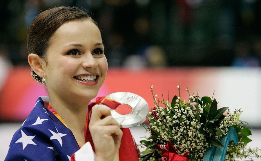 Sasha Cohen holding up her medal and flowers with an American flag wrapped around her 