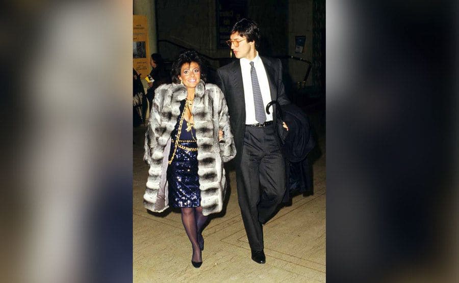 Maurizio walking with Reggiani who is wearing an elegant fur coat. 