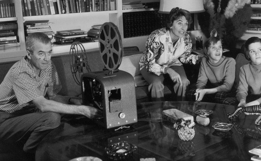 James Stewart operating a film projector while his wife and daughters sit by him in the living room waiting for the film to be ready 