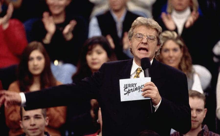 Jerry Springer with a microphone and cards speaking to guests on his show 