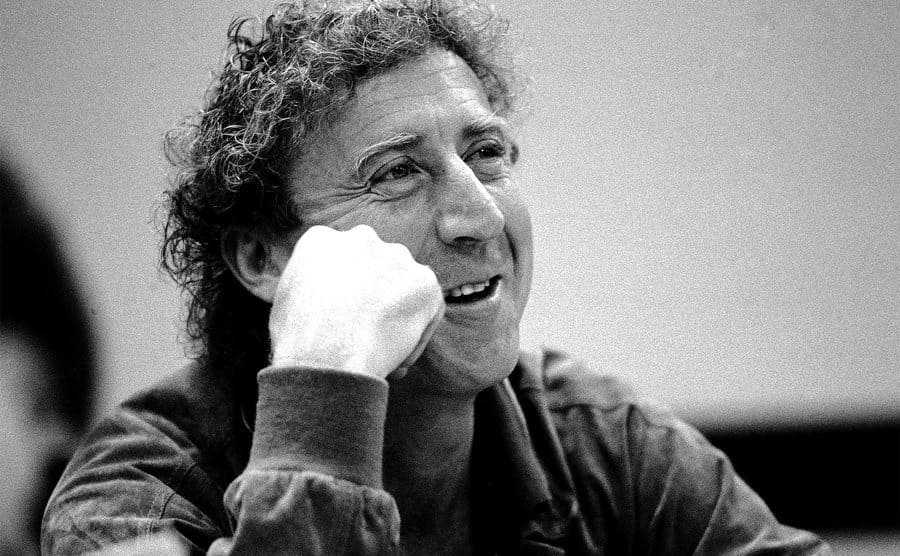 Gene Wilder is smiling as he rests his head on his hand. 
