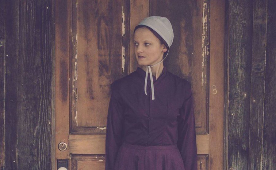 Emma Gingerich is leaning on the front door in Amish clothing and head covering. 