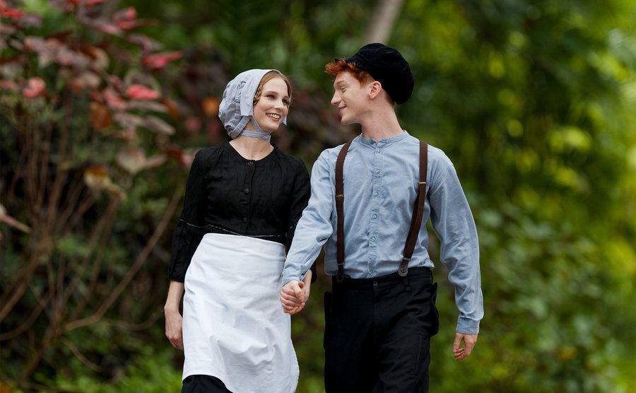 A young Amish couple are holding hands walking through the woods. 