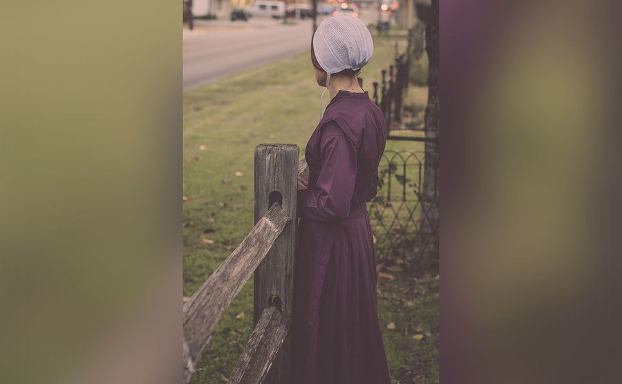 Emma stands by the fence in her Amish attire. 