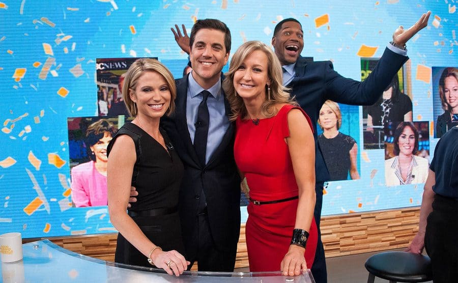 Amy Robach, Josh Elliott, and Lara Spencer posing for a photo with Michael Strahan photo bombing behind them. 